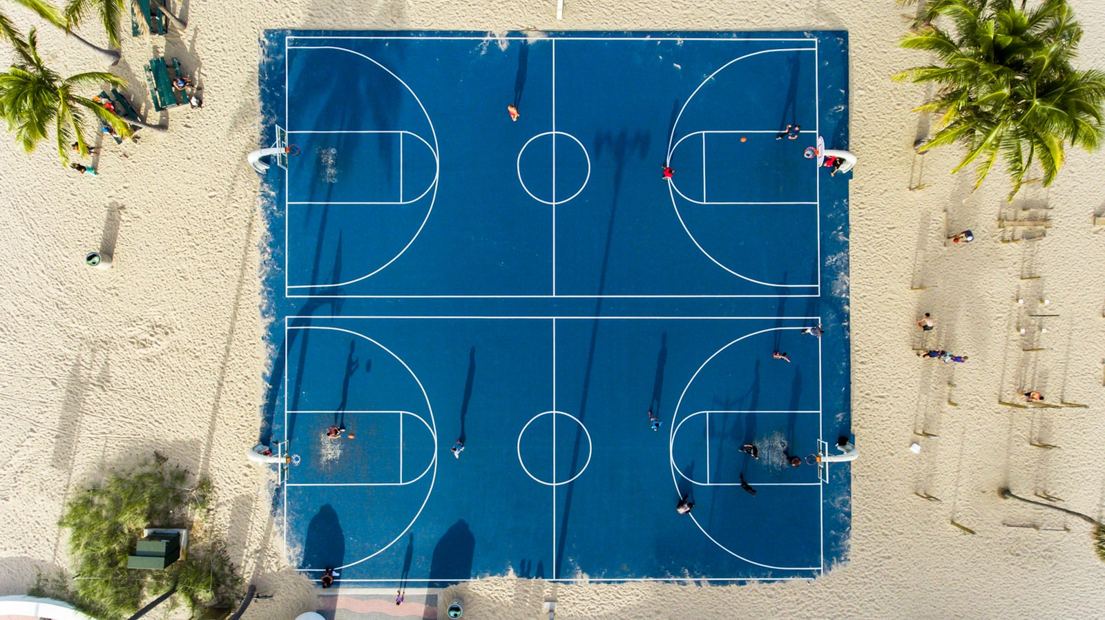 Veduta aerea di campi da basket blu sulla sabbia, circondati da palme, con partite in corso
